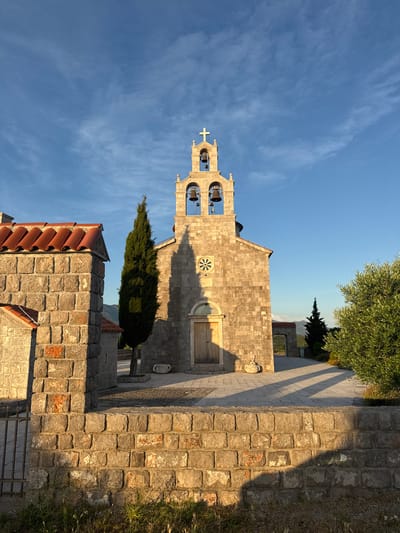 The image depicts a stone church in Kovačko Polje, Montenegro, bathed in the warm light of late afternoon or early morning. The sky is a clear, bright blue with wispy white clouds. In the foreground, a low stone wall with a red-tiled structure on its left separates the viewer from the church complex. The wall is constructed of rough-hewn gray stones with visible mortar. A metal gate is partially visible on the left side of the wall.
Dominating the center of the image is the stone church, constructed from large, rectangular gray stones. It features a prominent bell tower with three bells and a cross at its apex. The bell tower has arched openings and decorative stone carvings. The main facade of the church has a large wooden door with intricate carvings and a circular decorative element above it. A tall, slender cypress tree stands to the left of the church, casting a long shadow across the paved courtyard. To the right of the church, an olive tree with lush green foliage adds to the Mediterranean feel.
In the background, through an arched gateway, more of the church complex is visible, along with hints of rolling hills or mountains under the blue sky. The overall impression is one of tranquility and rustic beauty, characteristic of a Montenegrin village setting.