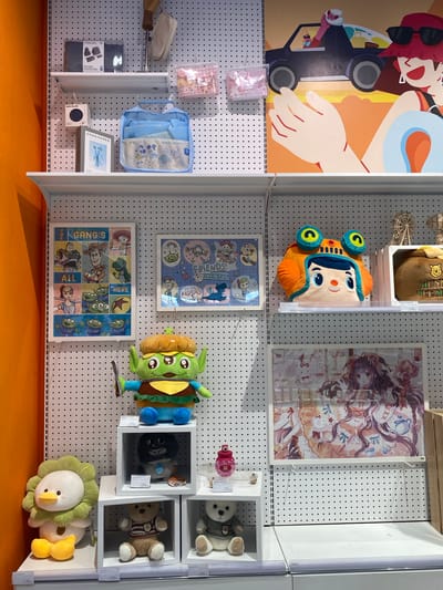 The image depicts a retail display for plush toys and merchandise, likely within a department store or specialty shop. The display features a pegboard wall with shelves, showcasing a variety of items. On the top shelf, there are travel bags and small pouches with cartoon characters. The middle shelf displays a "Toy Story" themed poster and a large plush character, along with a cartoonish character head pillow. Below this, more plush toys are arranged on cube shelves. The bottom shelf and foreground show additional plush animals, including a duck and bears, and a framed anime-style print. The overall impression is a colorful and whimsical collection of goods targeted at a younger audience or fans of popular franchises. The context of North Jakarta, Indonesia, suggests this scene is within a shopping mall or retail area in that region.