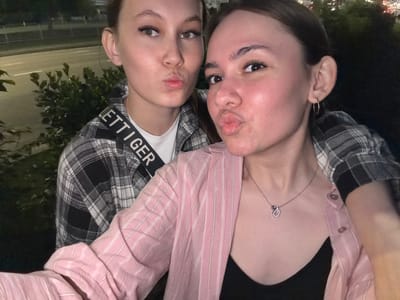 Two young women are taking a selfie outdoors at night. The woman in the foreground is wearing a pink striped shirt over a black top and a necklace. The woman behind her is wearing a plaid shirt. Both women are pouting their lips. The background shows a street with cars and trees. The location is the city of Naberezhnye Chelny, Russia.