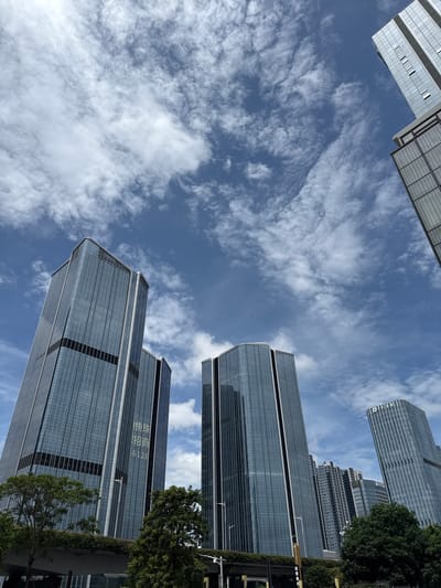 This is a low-angle, wide-shot photograph of modern skyscrapers in the Huangpu District, China, under a partly cloudy blue sky. The buildings are tall, with reflective glass facades, and some feature distinctive architectural elements like rounded corners and vertical accents. In the foreground, lush green trees and foliage obscure the lower portions of the buildings and a concrete overpass or bridge structure. On the facade of one of the central buildings, Chinese characters and numbers are visible, including "招商" and "6128". The sky is filled with wispy, white clouds that create a dynamic contrast with the deep blue sky. The image captures a typical urban landscape of a developed city, emphasizing the scale and modernity of its architecture against a natural backdrop of sky and vegetation.