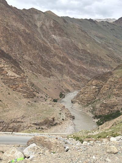 An overcast day in Reru, India, showcases a majestic, rugged mountain valley. A wide, muddy river flows through the center, carving its path between steep, rocky slopes. The mountains, predominantly brown and tan, are textured with fissures and scree, with sparse patches of dry grass clinging to the inclines. In the foreground, a paved road runs along the edge of the valley, bordered by a metal guardrail and scattered rocks. A bright green plastic bottle lies on the ground near the road, alongside other debris. Power lines are strung across the valley, suspended between poles on the mountainsides. The overall scene is one of raw, natural beauty, a testament to the powerful forces of erosion and geological formation.