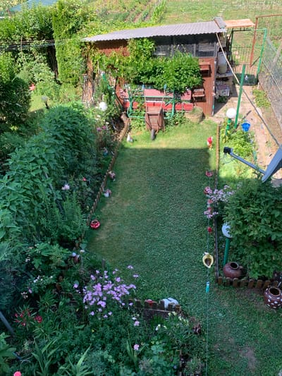 This is an aerial view of a backyard garden in Chernomorets, Bulgaria. The garden features a small wooden shed with a corrugated metal roof, a wheelbarrow, various potted plants, and a patch of grass. A wooden fence runs along the perimeter of the garden, separating it from other areas. Several decorative items, including ceramic pots and garden gnomes, are placed throughout the space. The backyard is surrounded by lush greenery and trees.