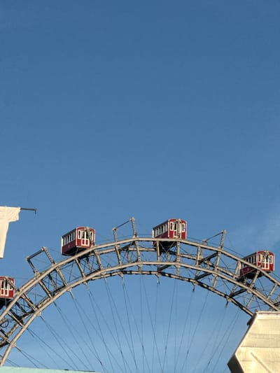 The image shows a section of the Wiener Riesenrad, a Ferris wheel located in the Prater amusement park in Vienna, Austria. The Ferris wheel is characterized by its distinctive red gondolas, several of which are visible against a clear blue sky. The intricate metal structure of the wheel is also apparent, with its spokes and supporting beams. In the lower right corner of the image, a portion of a light-colored building or structure is visible, likely part of the amusement park infrastructure. There are no people or ongoing actions depicted in the image, presenting a static view of the landmark.