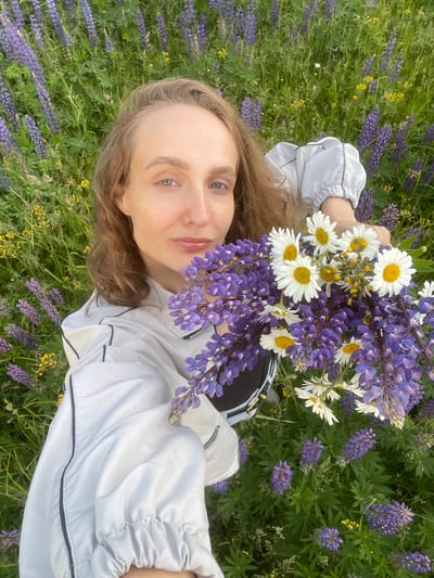 A young woman is taking a selfie in a field of lupines and daisies. She is holding a bouquet of lupines and daisies. The lupines are purple and the daisies are white with yellow centers. The woman is wearing a white jacket with black trim. The background is a blur of green foliage and purple lupines. The image appears to have been taken during the day in a rural area.