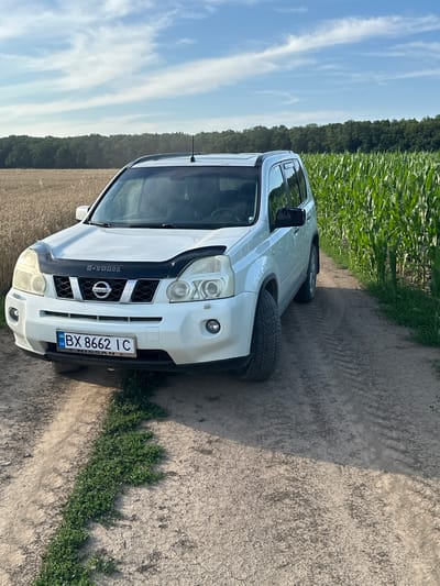 A white Nissan X-Trail is parked on a dirt road in a rural area. The car is facing the camera, with its front bumper and grille clearly visible. The license plate reads "BX 8662 IC". To the left of the car is a field of ripe wheat, and to the right is a field of tall green corn. In the background, a dense line of trees forms the horizon under a partly cloudy blue sky. The scene appears to be located in Slobidka-Zalisetska, Ukraine. There are no people or animals visible in the image. The overall environment suggests a sunny day in the countryside.