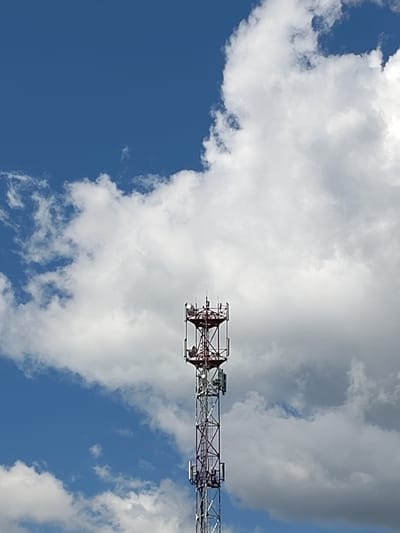 The image shows a tall telecommunications tower set against a bright blue sky filled with fluffy white clouds. The tower is a lattice structure with a red-painted upper section and a silver-painted lower section. Several antennas and equipment boxes are visible attached to the tower. The sky suggests it is daytime, likely with fair weather, as indicated by the clear blue sky and bright white clouds. The scene is outdoors, and the presence of a telecommunications tower implies a setting that requires connectivity, possibly in a semi-urban or rural area. No people are visible in the image. There is no visible text in the image. The location context provided is Ходаево, Russia.