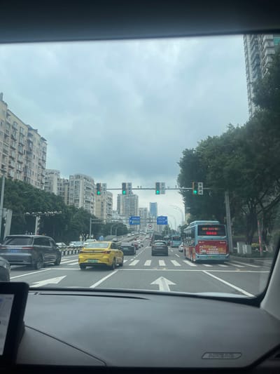 The image depicts a street scene from inside a vehicle, looking forward. The setting is an urban environment, likely in China, given the presence of Chinese text on traffic signs and vehicles. The sky is overcast, suggesting cloudy weather.

In the foreground, the dashboard of the vehicle is visible, including a navigation screen displaying the time "11:26" and what appears to be a map. The road ahead is multi-laned, with several cars and a bus. Lane markings are clearly visible, including a large white arrow indicating a forward direction.

On the left side of the road, there are tall residential buildings with numerous windows and air conditioning units. Trees line the side of the road, partially obscuring the buildings. On the right side of the road, more trees are present, as well as a tall modern building in the background.

Traffic signals are visible, with multiple sets showing green lights, indicating that traffic is allowed to proceed in those directions. Several traffic signs with Chinese characters are posted on poles. One sign, in the distance on the right, includes the text "重庆公交," which translates to "Chongqing Bus."

Vehicles on the road include a grey SUV on the left, a yellow sedan in the middle, and a blue and red bus on the right. The yellow sedan has a license plate that reads "川AZ0705." The bus has Chinese text on its rear, including "重庆公交," and an advertisement featuring the text "每天仅需1元" ("Only 1 Yuan per day").

The overall impression is a busy urban intersection during daylight hours under a cloudy sky. The location is identified as Longxi Subdistrict, China.