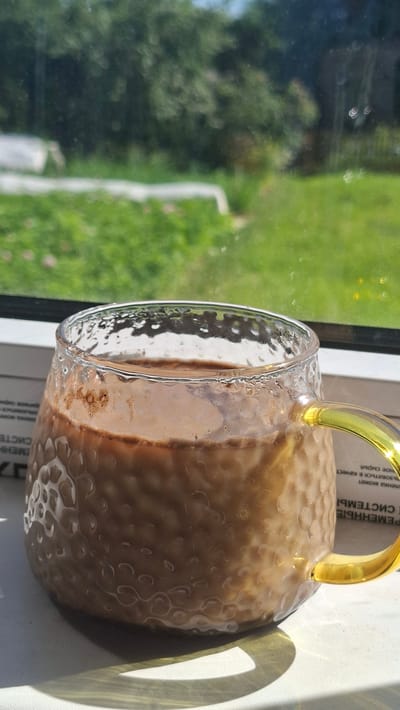 A close-up shot shows a glass mug filled with a light brown beverage, possibly coffee or hot chocolate, sitting on a white surface. The mug has a textured, hammered glass design and a bright yellow handle. The beverage has a frothy layer on top and appears to be warm, with condensation visible on the glass. In the background, through a window, lush green grass and trees are visible, suggesting an outdoor setting. The sunlight casts a distinct shadow of the mug onto the surface. The scene is set near a window, with some printed text visible beneath the mug on a white paper or book. The location is identified as Забалоцкі сельскі Савет, Belarus.