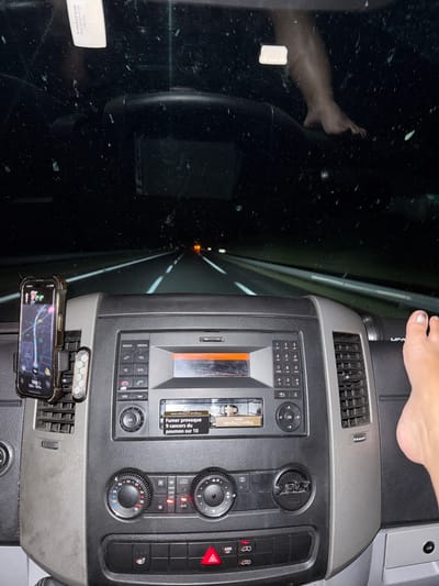 The image shows the interior of a vehicle at night, presumably driving on a highway. The dashboard is visible in the foreground, with a smartphone mounted on the left displaying a navigation app. The radio and climate controls are centrally located. A cigarette warning label is visible on the radio. The windshield shows streaks and dirt. In the upper right of the image, a person's bare foot is resting on the dashboard. The road ahead is dark with lane markers illuminated by the headlights. The overall impression is of a nighttime drive through a rural or suburban area, possibly near Castelnaudary, France, given the text on the cigarette warning which is in French.