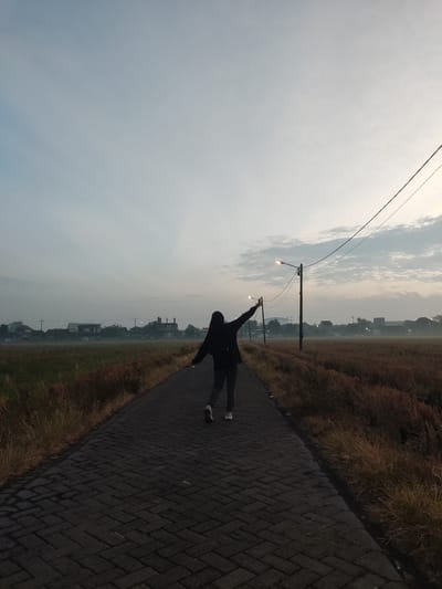 A person wearing a black hoodie and grey pants stands on a paved road with their back to the camera, facing away from the viewer. Their left arm is raised, and their right arm is extended to the side. The road is lined with dry, golden grass. In the distance, a few houses and buildings are visible through a light haze, with power lines and streetlights crossing the sky. The sky is pale blue with some scattered clouds. The scene appears to be in a rural or semi-rural area, possibly near agricultural fields, consistent with locations in Banjarkemantren, Indonesia, during dawn or dusk.