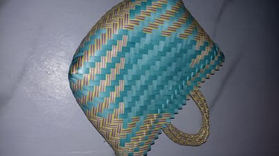 A woven basket with a teal body and yellow and purple accents sits on a gray, textured surface. The basket has a handle on the right side. The lighting creates a shadow on the left side of the basket, obscuring some of the details. The image is taken from a high angle.