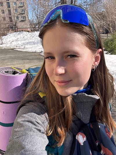 A young person is posing outdoors with a rolled-up purple yoga mat visible in the foreground, and possibly wearing a gray jacket and blue goggles. Behind the person is a building and some patches of snow. The location may be Berezniki, Russia.