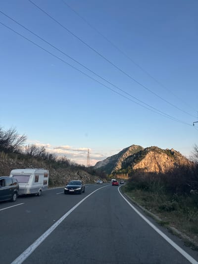 The image captures a scenic road view in Mišići, Montenegro, with a clear blue sky and rugged mountains in the background. A caravan is being towed by a car on the right side of the road, heading towards the mountains. Several other vehicles are visible on the road, creating a sense of traffic flow. The sun casts a warm glow on the mountains, highlighting their rocky textures and sparse vegetation. Overhead power lines traverse the sky, indicating the presence of infrastructure in the otherwise natural landscape. The overall scene suggests a late afternoon or early evening setting, with the low sun illuminating the peaks.