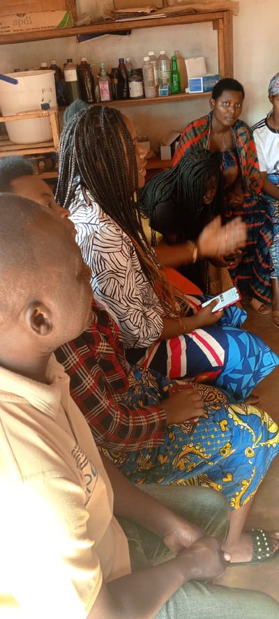 This indoor image from Ngalula, Democratic Republic of the Congo, shows a group of several individuals, predominantly women, seated closely together in what appears to be a community or functional room. They are engaged in a meeting, discussion, or training session, with most looking towards an unseen central point.
The subjects are dressed in various patterns, including vibrant African wax prints, a black-and-white zebra-like design, and plaid. Many have braided hairstyles. In the foreground, the side profile of a man is visible, and next to him, a woman in a black-and-white patterned top holds a red smartphone. Another woman further back is actively gesturing with her hand while also holding a phone.
The background features simple wooden shelves lined with an assortment of bottles (some dark, some clear), a white bucket, and a white basin. Several boxes are also on the shelves. Visible text includes "apdl" in green on a cardboard box on the top shelf, accompanied by the phrases "WITH HEALTH" and "WITH US. LIFE ALWAYS GETS BETTER." Another box on a lower shelf is labeled "Surgical Mask," hinting at a potential medical or health-related context for the supplies and the gathering. On one of the smartphones, the text "REZ" is visible on the screen.
The scene is lit by natural light, suggesting it is daytime. The general mood appears attentive and engaged.