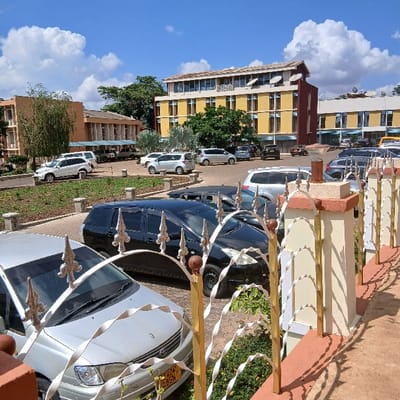 The image displays an outdoor scene, rotated 90 degrees counter-clockwise from its likely original orientation, showing a row of parked cars alongside a street or driveway, with buildings and a clear sky in the background.
In the immediate foreground, on the right side of the image, there is a decorative fence or railing. It features white and gold-colored sections with intricate, swirling patterns, supported by reddish-brown posts that have lighter, cream-colored accents. Some spherical finials are visible on the fence posts.
Behind the fence, a paved area, which appears to be a parking lane or driveway made of light brown interlocking pavers, stretches into the distance towards the left background. Numerous vehicles are parked along this lane. The closest visible car, partially obscured by the fence, is a silver or light grey sedan. A yellow license plate is clearly visible on its front, bearing the characters "NBB 823". Behind it, a black minivan or SUV is parked, followed by another dark-colored car, and then a white SUV or minivan. Further down the lane, several more white sedans or small SUVs, a black car, and a yellow van or small bus are visible.
To the left of the parking lane, a strip of green grass and various plants, including some small trees and palm trees, separate the cars from the buildings.
In the mid-ground and background, several buildings are visible. The most prominent building, on the left side of the image, is a multi-story structure painted predominantly yellow with brown trim and a reddish-brown tiled roof. It has numerous rectangular windows, and several satellite dishes are mounted on its rooftop. Beyond this building and further into the background, other structures of varying colors (browns, light orange) are interspersed among green foliage.
The sky above is bright blue with scattered, fluffy white cumulus clouds, indicating clear and sunny weather. The bright illumination suggests it is daytime, likely mid-morning or early afternoon, with visible shadows cast by the objects. No specific activities are ongoing; the scene depicts a static arrangement of parked vehicles and buildings.