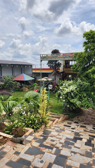 This is an outdoor shot of a garden area with a play structure and a two-story building, taken from an elevated perspective looking down a stone staircase. The garden features lush green grass, various plants, flowers, and palm trees. A colorful umbrella-shaped canopy covers a play area with a slide. To the right, a large tree with white flowers dominates the foreground. The building has multiple levels, with balconies and an exterior decorated with red, yellow, and green stripes. A windmill decoration is visible on the side of the building. The sky is filled with fluffy white clouds. The scene appears to be in a public space, possibly a cafe or park, in the city of Ia Hru, Vietnam. There are two individuals visible in the background, near the building, appearing to be casually walking.