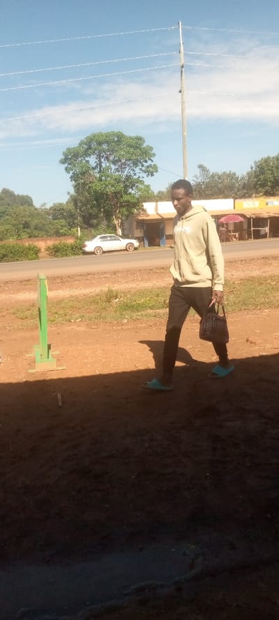 The image shows a young man walking on a dirt road in Kivaywa, Kenya (0.56, 34.806). He is wearing a light brown hoodie, black pants, and blue flip-flops. He is carrying a small, dark brown handbag. There is a white car parked on the road behind him. The man is walking away from a tall wooden pole with multiple electric wires attached. There are trees and foliage in the background. The ground is dirt, with some grass and foliage growing in areas. The sky is clear and blue. The image is taken from a slightly elevated angle.