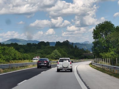 This is a ground-level, eye-level shot of two cars driving on a highway in Osikovitsa, Bulgaria. The day is overcast with scattered white clouds in a light blue sky. The scene is framed by lush green trees on either side of the highway, with a backdrop of rolling, forested hills and mountains in the distance.
The primary subjects are a dark BMW SUV in the left lane and a white Toyota RAV4 SUV in the right lane, both traveling away from the camera. The BMW has license plate "CB7979PP". The Toyota has a spare tire mounted on its rear hatch and license plate "BT 1852KH". A white truck is visible in the distance on the left shoulder of the highway. A concrete overpass with guardrails is visible in the middle distance on the right side of the highway, over which the road curves to the right. Metal guardrails with reflective markers line the sides of the highway.
The activity is a journey or travel on a highway. The time of day appears to be daytime, likely late morning or afternoon, given the brightness of the sky and the shadows. The weather is fair, with clouds suggesting a possibility of rain but no active precipitation is visible. The presence of a highway, guardrails, and distant mountains places the location in a scenic, possibly rural or semi-rural area. No people are directly visible within the vehicles.
