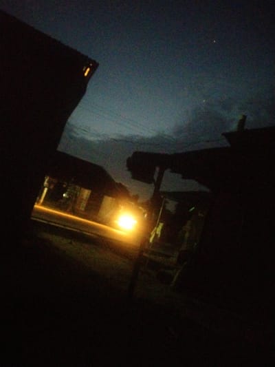 The image appears to be taken at night in what may be Uyo, Nigeria. The scene is mostly dark with silhouettes of buildings on either side. A street runs between them, and a bright light, possibly from a car's headlights, illuminates part of the road and nearby structures. Two small points of light in the sky, possibly stars, are visible through gaps in the buildings. There are no clearly visible people. The environment is urban or suburban, with buildings and a road. The photograph seems to be taken from a low angle.
