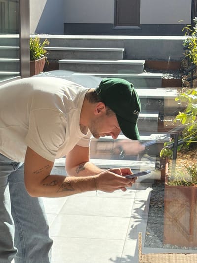 The scene is located in Marche-lez-Écaussinnes, Belgium. A male individual, viewed in profile from the mid-torso up, leans forward slightly while holding a light-colored smartphone horizontally with both hands. He is wearing a dark green baseball cap with light embroidery, a plain white short-sleeved t-shirt, and light blue jeans. His left forearm displays tattoos including text, a bottle, and a mountain range, while his right forearm has a butterfly tattoo. A thin chain bracelet is visible on his left wrist. The image is captured through a reflective glass window, showing an outdoor area beyond. This area features light-colored square patio tiles, a series of light grey concrete or stone steps leading upwards, and a building facade with a dark window or door frame. Planters with green foliage are present; one in dark wood on the left and another in a rusted metal material on the right, surrounded by dark gravel. Strong sunlight illuminates the scene, casting distinct shadows.