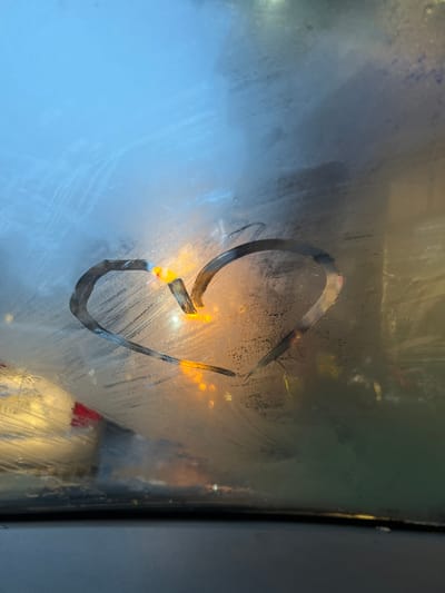 The image displays a heart shape manually traced onto the interior condensation of a vehicle's windshield. The cleared pathways of the heart reveal blurred external artificial light sources, predominantly amber and white, indicating an urban environment under low-light conditions. A dark vehicle dashboard is visible at the bottom of the frame. No individuals are depicted, nor are there any discernible landmarks or identifying features within the image to determine a specific location in Barza Municipality, Syria.