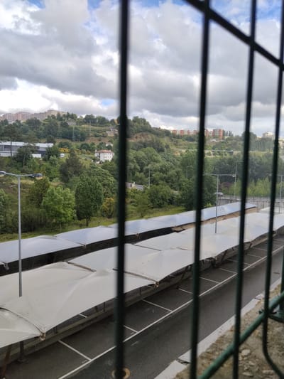A daytime shot through a metal barred window shows a covered parking lot in Vila Real, Portugal. The parking lot features multiple rows of awning-covered spaces. Behind the parking lot, a hill rises, densely vegetated with trees and interspersed with lampposts. Buildings are visible on the hilltop, some appearing as apartment blocks. The sky is overcast with patches of blue. The focus is on the scene outside the window.