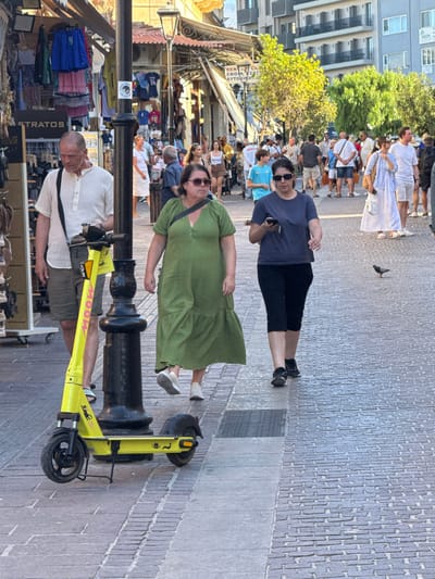This image captures a bustling street scene in Chania, Greece, likely in the old town. A man in a light-colored shirt and shorts walks past a black lamppost, with a bright yellow electric scooter parked next to it. Two women are walking together in the foreground; one in a green dress with a crossbody bag, and the other in a dark blue t-shirt and capri pants, holding a phone. The street is paved with cobblestones and lined with shops displaying clothing and merchandise. In the background, more people are walking, and buildings with balconies are visible. The lighting suggests it is daytime, possibly late afternoon, with sunlight casting shadows. A pigeon is seen on the ground near the walking women.