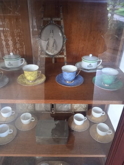 The image shows a glass-fronted display case containing a collection of porcelain teacups and saucers, framed photographs, and other decorative objects. The display case is likely located in a museum or private residence in Surakarta, Indonesia, given the coordinates provided (latitude: -7.566, longitude: 110.823). The teacups are arranged on two levels within the case, with some saucers overlapping. There is a small, rectangular box on the lower level of the display. The top level features a picture of a woman in a long white dress with an ornate frame. The frame is made of wood and is embellished with gold trim and jewels. The woman in the picture appears to be seated and her face is partially obscured by her hair. The image is a testament to the beauty and artistry of the past.