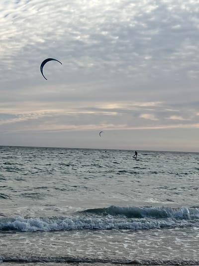 The image depicts a cloudy day at the beach in Tarifa, Spain. The sea is choppy with moderate waves breaking near the shore. In the distance, two individuals are kite surfing. One kite surfer, a silhouette against the water, is visible on a board, appearing to be actively engaged in the sport. Another kite is seen further out in the water, also appearing to be in use. The sky is overcast with textured clouds, obscuring the sun but allowing some ambient light. The foreground shows the sandy shore with the incoming waves lapping at the beach.