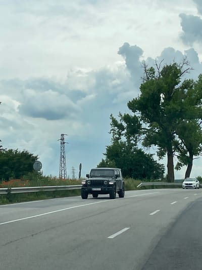 The image shows a road with two vehicles traveling on it, under a sky filled with clouds. In the foreground, a dark-colored Hummer is seen. Further down the road, a white car is also visible. The road has lane markers, and on the sides, there is greenery, including trees. In the background, there is a metal electrical transmission tower. Veselina, Bulgaria is not explicitly visible, but the scene depicts a road and environment consistent with a rural area that could be found in Veselina.