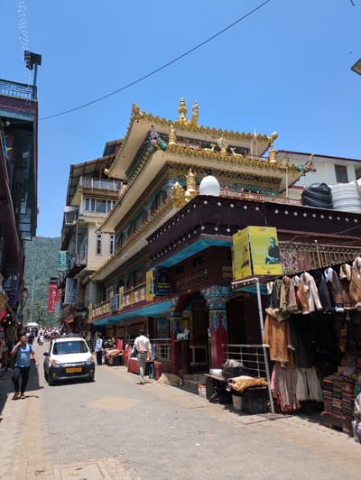 The image depicts a street scene in Dharamshala, India, with a clear blue sky overhead. The street is paved with stone blocks and lined with buildings, some of which feature traditional Tibetan architectural elements. The most prominent building on the right is adorned with intricate gold decorations on its roof and colorful murals. In front of this building, several shops display various items for sale, including clothing racks filled with jackets and piles of textiles.

In the street, a white car is visible, with its license plate reading "HP01 R 4497". Several people are present: a man in a blue t-shirt and dark pants walks towards the viewer, talking on his phone. Another man in a white shirt and light-colored pants walks away from the viewer further down the street. A woman in a light blue shirt and dark pants stands near the white car, and other individuals are seen in the background, suggesting a bustling marketplace. A small, light-colored dog is lying on the ground to the right of the street. The buildings in the background are multi-story structures with balconies and windows, and a forested hillside is visible in the distance. The overall impression is that of a vibrant street in a town or city, likely a tourist area due to the shops selling goods.
