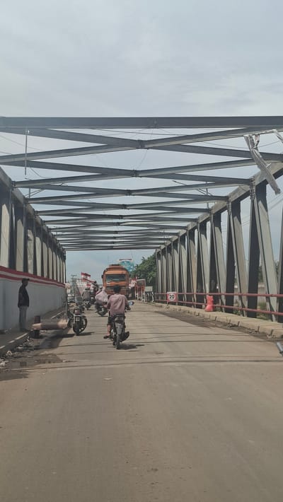 A traffic scene unfolds on a bridge in Kulukuta, Indonesia, under a bright, clear sky, suggesting daytime. The perspective is from a vehicle moving across the bridge, offering a view through the steel trusses of a large, industrial-looking structure. The bridge's metal framework dominates the left side of the frame, creating a repeating geometric pattern against the pale sky.

On the right side of the bridge is the road, paved with asphalt and showing signs of wear. Traffic is present, consisting mainly of motorcycles and a small orange vehicle that resembles a tuk-tuk or a mini-bus. Several people are visible riding the motorcycles, some with passengers. The figures are indistinct, but their presence indicates normal daily activity. A red sign with "20" is affixed to a railing along the side of the bridge, likely indicating a speed limit. Further down the road, other vehicles and some distant buildings or structures can be seen. The overall atmosphere is that of a busy, functional bridge serving as a route for local transportation.
