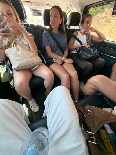 The image depicts four individuals in the back seat of a car, seemingly on a road trip. The perspective is from the interior of the car, looking towards the front.

In the foreground, a young woman with blonde hair is looking down at her phone, which she is holding in her left hand. Her right hand is resting on a light-colored woven purse. She is wearing a white t-shirt.

Behind her, another young woman with dark hair is wearing a blue t-shirt and a seatbelt. She is looking towards the camera with a surprised or confused expression. A black backpack is visible behind her seat.

Further back, partially obscured, are the legs of at least two other individuals. One person appears to be wearing dark pants, and another has tan legs and is wearing white sneakers. A brown suede bag with gold buckles is also visible. A water bottle with a blue cap is placed on the white fabric in the foreground, suggesting a sunny day and the need for hydration.

The setting appears to be a car's interior, with car seats and windows visible. The exterior visible through the rear window suggests a sunny day with dry, possibly rural or coastal scenery. The time of day is likely daytime, given the lighting. The overall mood seems relaxed and casual, consistent with a road trip. No visible text is discernible in the image. The location context of Fréjus, France, hints at a Mediterranean setting.