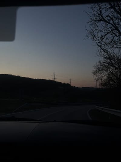 The image captures a twilight scene in Stremtsi, Bulgaria, viewed from inside a vehicle looking through its windshield. The lower approximately one-quarter of the frame shows the dark dashboard. Above this, a paved road with white lane markings curves gradually to the right, with a metal guardrail present on the right side of the road. In the midground, dark, densely vegetated hills form the horizon line. Several large electricity pylons carrying power lines are visible across the hills and against the sky. On the far right, a cluster of bare, leafless tree branches is silhouetted. The sky exhibits a gradient, transitioning from a darker blue-grey at the top to a lighter, faint orange-pink hue near the horizon, consistent with dusk. No individuals or active interactions are visible.