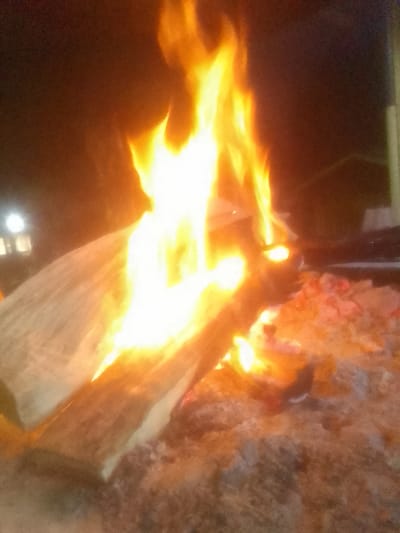 A vibrant bonfire dominates the scene, featuring tall flames reaching upwards. The fire engulfs large pieces of wood and glowing embers rest beneath. Light illuminates the dark foreground and a building is partially visible in the background. The overall impression is warmth and a sense of being outdoors at night.