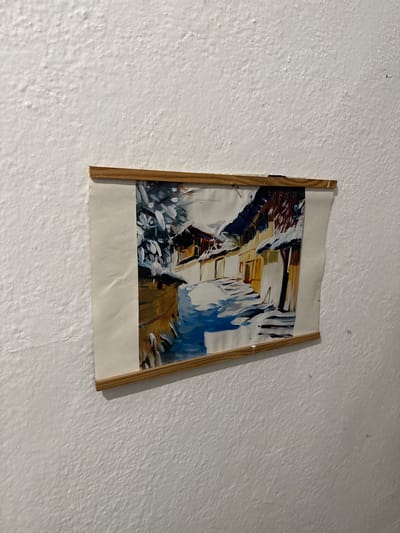 A painting of a snowy village scene is displayed on a white, textured wall. The painting depicts snow-covered buildings with warm-toned facades lining a pathway. The sky appears to be a muted blue and white, suggesting a winter day. The artwork is held by a simple wooden frame that is attached to the wall. The overall impression is one of a cozy, rustic setting.