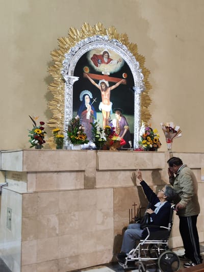A full shot depicts two people in what appears to be a church in Tacna, Peru, in front of a large Catholic altarpiece. On the left, a woman sits in a wheelchair, facing the altarpiece. Her right arm is raised, her hand pointing upwards. She wears glasses, a dark blue jacket over a striped shirt, and grey pants. Next to her and to her right stands a man with short, dark hair. He wears a light tan jacket and black athletic pants with white stripes. His right hand is raised to his forehead as if in prayer or reflection. The altarpiece behind them dominates the scene. It features a painting of the crucifixion of Jesus, flanked by Mary and another figure. Above Jesus, the painting depicts God the Father with a dove. The painting is framed by a silver archway adorned with gold rays emanating outwards. In front of the painting, on a marble-like platform, are several bouquets of flowers. The wall behind the altarpiece is a light beige color. To the left of the scene, a marble structure is visible. The floor is grey. Several crosses are painted on the wall to the right of the man and woman.