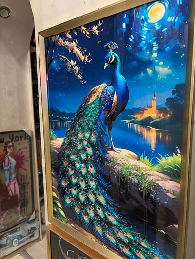 The image shows a framed artwork of a peacock in a night scene. The peacock is depicted with its tail feathers fanned out, adorned with sparkling embellishments. It stands on a rocky outcrop overlooking a body of water, with a castle or church illuminated in the background under a moonlit sky. The artwork is framed in gold. To the left of the peacock artwork, there are other framed pictures, one of which appears to be a fashion illustration with a woman holding a handbag and another with a vintage car. The setting appears to be an indoor display, possibly a gallery or shop, with the wall texture and lighting suggesting an interior environment. There is no information about the city of Sandanski, Bulgaria in the image.