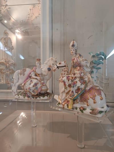This media file features two ornate porcelain figurines displayed in a glass-enclosed cabinet, likely in a museum setting. The figurines depict regal figures on horseback, adorned with elaborate floral patterns and gold accents.
The figurine on the left shows a woman in a crown, seated on a white horse. She wears a richly decorated gown and holds a scepter. A globe rests at the horse's feet, suggesting themes of power or exploration. The horse itself has a patterned saddle blanket.
The figurine on the right features a figure, possibly also a woman given the attire, seated on a more dynamic, perhaps mythical, creature that seems to emerge from stylized water or foliage. This figure also wears a decorative headdress and flowing garments, embellished with floral motifs and gold trim. The creature has a flowing mane or tail rendered in turquoise and white.
Both figurines are presented on white, decorative bases depicting foliage and flowers, and are elevated on clear acrylic stands within the display case. The background is blurred, showing the interior of the cabinet and possibly other museum exhibits, with reflections on the glass. The overall impression is one of historical artistry and craftsmanship, with the polished surfaces and bright lighting suggesting an indoor exhibition space. The location context provided is Colorno, Italy.