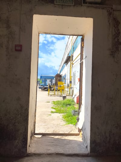 This is a photograph taken from inside a dark, industrial-looking building, looking out through a large, rectangular opening. The opening reveals a brightly lit outdoor scene. The interior walls are rough and textured, with some visible damage and staining. On the left interior wall, there is a red fire alarm. Above the opening, there is a sign with Russian text, which appears to say "ВЫХОД" (VYKHOD), meaning "EXIT".
The outdoor scene is bathed in daylight, suggesting it is daytime. The sky is a vibrant blue with scattered white clouds, indicating fair weather. Outside the building, there is a paved or dirt ground area, with some patches of green grass. In the distance, a long, white industrial building with blue and yellow trim is visible. Various objects are present, including a blue truck, a yellow metal fence, and some yellow industrial equipment. The overall setting appears to be an industrial yard or a factory exterior. There are no people clearly visible in the frame, though the presence of a truck and industrial buildings suggests an active industrial area.