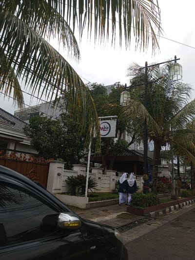 This image captures a street scene in Cianjur, Indonesia, under an overcast sky. The lower left quadrant is occupied by the front left portion of a black vehicle, specifically its hood and driver-side mirror, which exhibits an amber turn signal. Overhanging palm fronds frame the upper section of the image.
In the midground, a substantial building with a dark-tiled roof and white facade is partially visible behind an ornate brown gate and a decorative white concrete fence. The fence features recessed lighting and extends along a paved sidewalk. A white rectangular sign displaying "mbiz" in purple lettering with "marketplaces" in red underneath is mounted on a pole positioned between the fence and the sidewalk.
To the right of the sign, two individuals, appearing to be female students, walk together on the sidewalk, moving away from the viewer. They are dressed in dark blue tops, white long garments (skirts or trousers), and white head coverings. Adjacent to their path stands a black street lamp pole fitted with two ornate glass lanterns. This pole is situated next to a palm tree laden with visible coconuts. The scene is bordered on the right by an asphalt road with a black and white striped curb, separated from the sidewalk by a narrow strip of green landscaping and bushes. Dense green foliage, including various trees and shrubs, is prevalent throughout the background and midground.