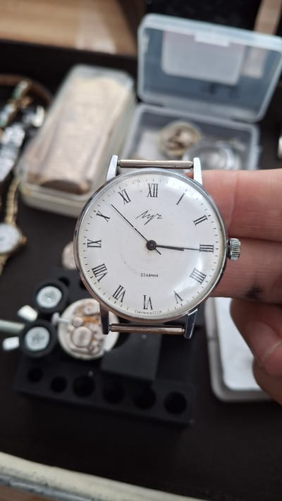 In Kempelevo, Russia, a close-up shot shows a vintage men's wristwatch being held by a person's fingers. The watch face is white with black Roman numerals and a small seconds sub-dial at the 6 o'clock position. The brand name "Луч" (Luch) is visible above the sub-dial. Below the sub-dial, "23 КАМНЯ" (23 Jewels) and "СДЕЛАНО В СССР" (Made in USSR) are inscribed. The watch has a silver-toned case and a metal strap. In the background, out of focus, are various small objects and a plastic container, suggesting a storage or collection setting.