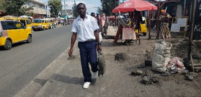 A man dressed in a light-colored shirt and dark pants walks on a sidewalk, holding a briefcase. The background reveals a bustling urban scene with yellow vehicles lining the street, suggesting a transportation hub. A red umbrella shades a vendor's stand on the right. The sidewalk is uneven, with a large bag and dark material scattered around. A woman with a child stands near a chalkboard, adding to the busy street life.