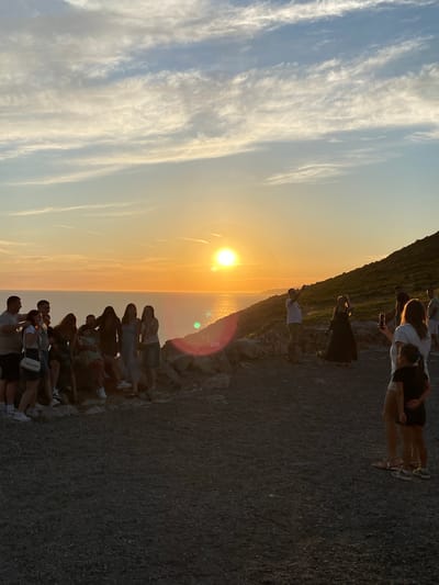 The image depicts a group of people gathered on a rocky terrain overlooking the sea at sunset. The sun is low on the horizon, casting a warm golden light across the scene and reflecting on the water. Some individuals are standing, while others are seated. Several people are holding up their phones, seemingly capturing the moment. A man in a white shirt and khaki shorts is standing and looking towards the sun. In the foreground, two young girls are visible, one with her arm around the other. The environment is characterized by rugged, rocky ground and a sloping hillside covered in vegetation in the background. The sky is a blend of orange, yellow, and blue, with wispy clouds. The location is identified as Kovačko Polje, Montenegro.
