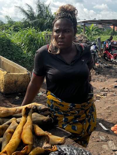 The image, taken in Obosi, Nigeria, shows a woman standing in what appears to be an outdoor market. She is wearing a black t-shirt and a yellow skirt with a blue and white patterned design. Her hair is styled in braids with blonde highlights. She is looking at the camera with a serious expression. In front of her, there is a pile of what looks like animal parts, possibly meat or offal, arranged on a yellow surface. The ground is dirt and there is debris scattered around. In the background, there are some structures, including what looks like a roofed area and some vegetation. A red motorcycle is also visible. The lighting suggests it is daytime. The overall scene appears to be a bustling, possibly informal, marketplace.