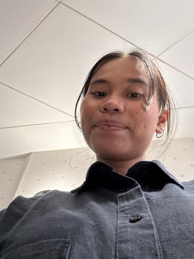 An overhead, slightly distorted, close-up, chest-up selfie of a young woman with medium-dark skin and long, dark brown hair. She is wearing a dark blue collared shirt and a hoop earring in her left ear. She has a small mole on her left cheek. The background consists of a white tiled ceiling and a wall with a light beige wallpaper featuring a subtle blue diamond pattern. The photo appears to have been taken indoors, possibly in a residential or office setting. There is no discernible information in the image to indicate a specific location within South Jakarta, Indonesia.