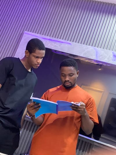 In a location identified as Igboefon, Nigeria, two adult males are observed standing indoors, focused on a shared object. The male on the right, wearing an orange short-sleeved t-shirt and displaying a beard, holds an open blue notebook or book with visible white pages, appearing to read or examine its contents. A gold-colored ring is present on his left ring finger. The male on the left, wearing a black short-sleeved t-shirt, is positioned slightly behind and to the left of the first individual, observing the same open notebook. The background consists of a vertically corrugated or ridged wall section, illuminated by purple ambient lighting, and a smooth, possibly reflective, horizontal surface also accented with purple light. The overall environment suggests a modern indoor setting.