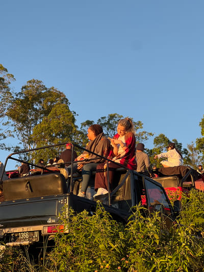 In Kintamani, Indonesia, a group of people are gathered atop two off-road vehicles, likely partaking in a sunrise tour, a popular activity in the area. The sky is a clear, vibrant blue, indicating fair weather. Lush green foliage surrounds the vehicles, suggesting a natural, possibly mountainous, setting. One vehicle, primarily black, has a visible license plate with the characters "F 1366 JV" and "03-27." A woman with blonde hair tied in a ponytail, wearing a red and white patterned shawl over a white top, is seated on the edge of this vehicle, looking down at her phone. To her left, a woman with dark curly hair is seated, wearing a gray blanket with a colorful pattern and teal leggings. Other individuals are also seated or standing on and around the vehicles, some with their backs to the camera. The lighting suggests it is either early morning or late afternoon, with the warm tones of the sunlight illuminating the scene.