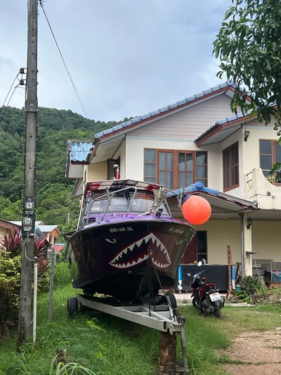 In the city of Choeng Thale, Thailand, a dark purple boat with a shark-mouth design on its hull is parked on a trailer in a grassy area. The boat is positioned in front of a two-story house with beige siding and blue-tiled roofs. An orange buoy is attached to the boat's railing. To the right of the boat, a red motorcycle is parked on a gravel path. A wooden utility pole with electrical equipment stands to the left of the boat, with green foliage and a forested hillside in the background. The sky is overcast.