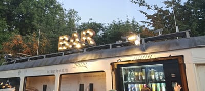 An outdoor scene at dusk features a white train car serving as a bar. A large, illuminated sign spelling "BAR" with incandescent bulbs is mounted on the roof of the train car, above a refrigerated display case filled with drinks. Text printed on the side of the train car includes "Don't forget the ice, baby" and menu items such as "APERITIVO SPRITZ &". A person's hand is visible near a keypad on the side of the refrigerated unit, suggesting a transaction is taking place. The background consists of green trees and a light blue sky. The scene appears to be at the Postlingbergbahn station in Linz, Austria.